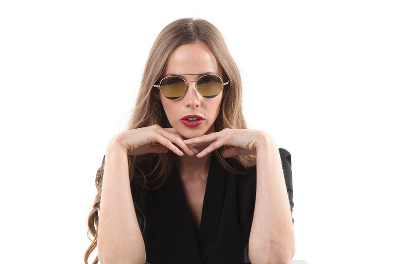 A woman wearing sunglasses and a black outfit posing with her hands under her chin against a white background.
