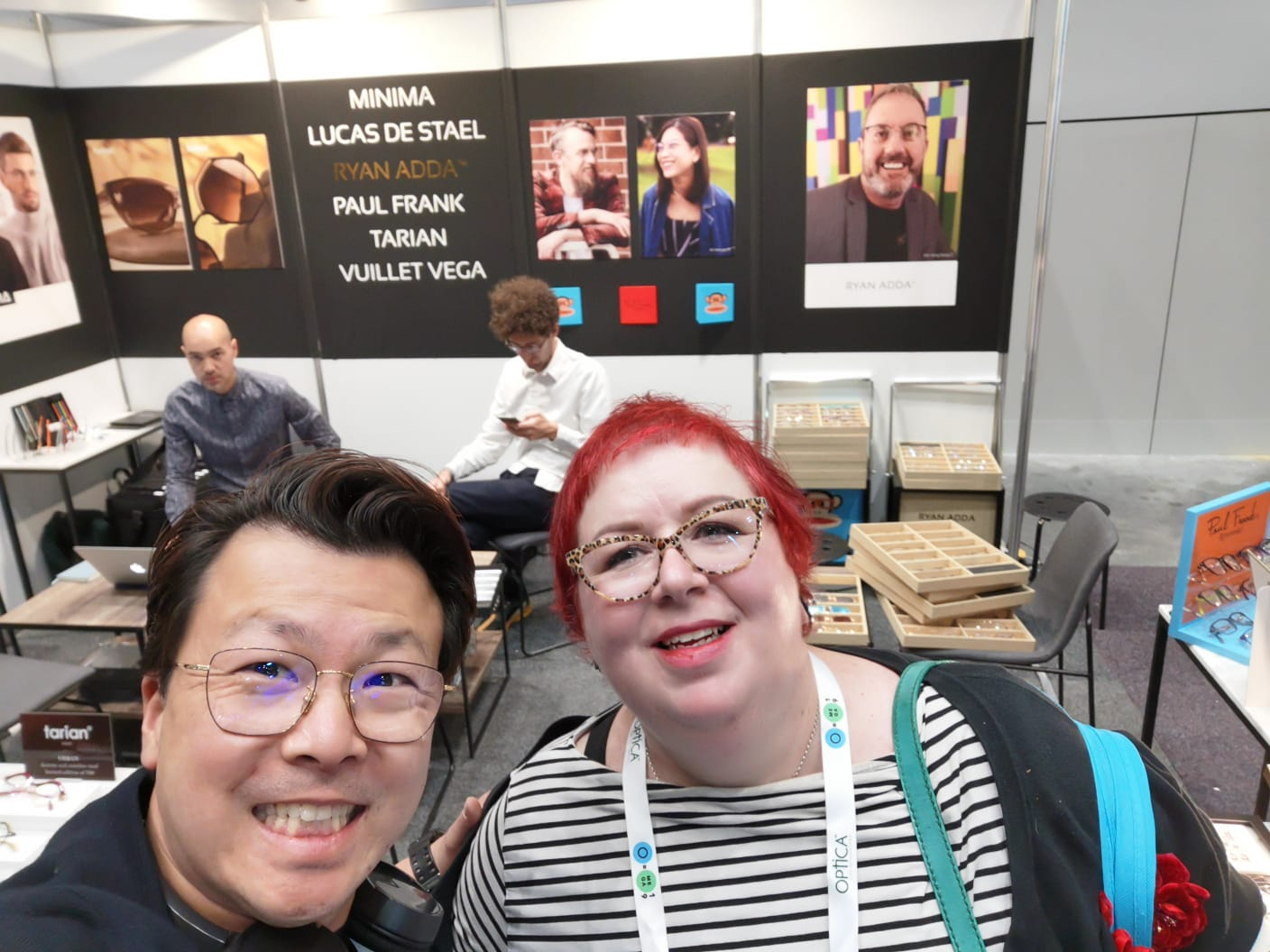 Two individuals are smiling for a selfie at a booth with promotional materials and people in the background at an event or conference.