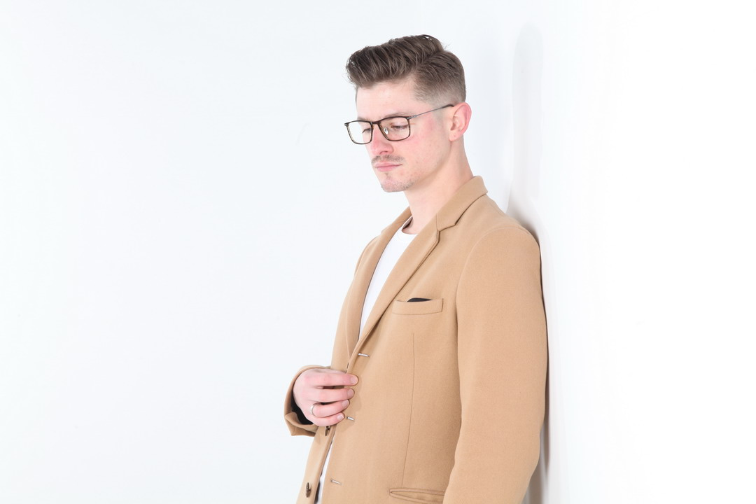 A man wearing glasses and a tan coat stands against a white background with a thoughtful expression, looking to the side.