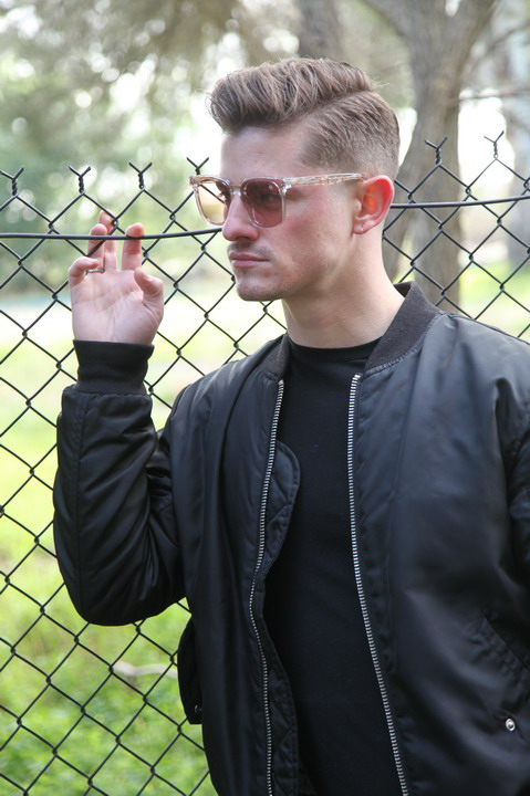 A man with sunglasses and a black jacket is standing by a chain-link fence in what appears to be a park.