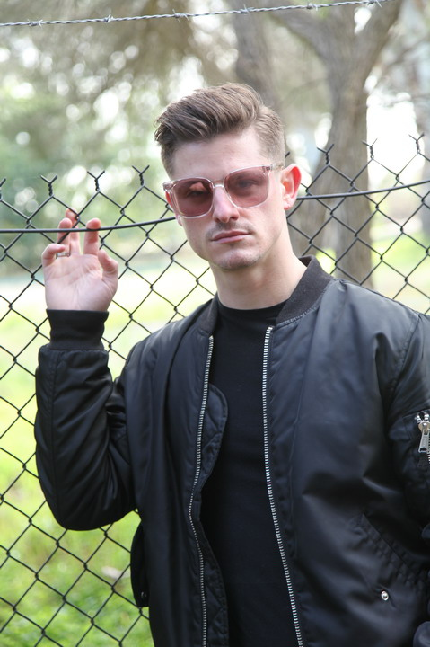 A man with sunglasses and a black jacket is posing in front of a chain-link fence.